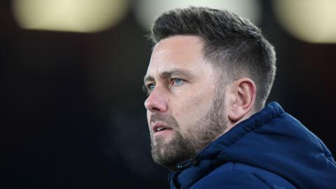 Oxford United manager Des Buckingham looking on at a football match, wearing a blue winter coat. He has short dark hair and a thin dark beard.