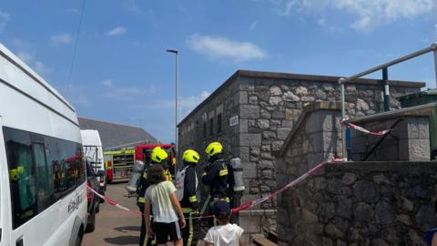 A fire truck in the distance and three crew members with florescent yellow helmets in the foreground, behind some fire service red and white tape,  which is blocking off the public toilets