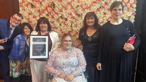 A group of five women and one man are in front of a flower wall. The woman in the middle is sat in a wheelchair and the woman to the left of her holds up a certificate.