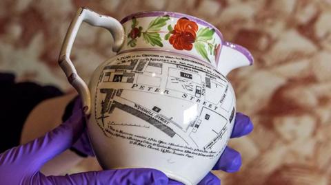 Close-up of a white rare commemorative jug of the Peterloo Massacre which features a map of the site when the 1819 tragedy unfolded. The lip of the jug has a red flowers with green leaves and a purple rim. Someone is holding the item with purple gloves.