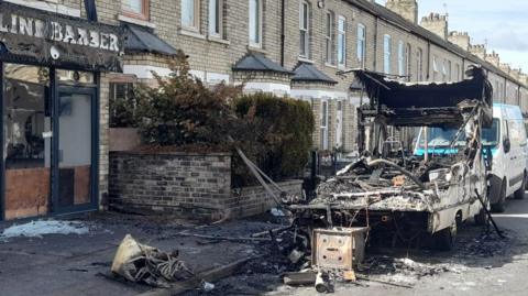 The image shows the aftermath of a vehicle fire on a residential street outside a row of terraced houses. The street is lined with parked vehicles. In the centre-right of the image is a severely burnt-out campervan. The vehicle is destroyed, with the metal frame exposed and blackened. 