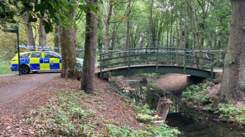 A yellow and blue police car can be seen parked on a path next to a dark green bridge which is curved across a stream of water in a woodland. A police white and blue striped cordon is placed from where the car is across to a tree on the other side of the bridge.