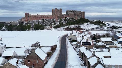 Bamburgh Castle rises above a snowy Bamburgh village. A winding narrow road leads towards the castle with the North Sea visible in the distance. 