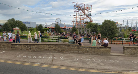 People walk around the periphery of the Skegness compass gardens.  Various rides provide the back drop and the shot is framed by festoon lighting.