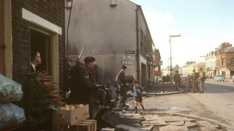 A photo from the Falls Road Belfast in the 1969s. It shows a street with British soldiers. A boy in a blue top and shorts are walking past them. A man in the left hand corner is looking outside an open door. There are also boxes of produce outside the door. 