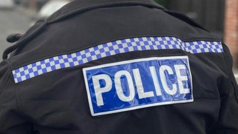 The back of a black police jacket with 'Police' written in white on a blue background. A strip of chequered blue and white squares runs across the top of the jacket.