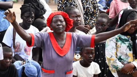 A crowd of Catholic women and children kneeling on the ground outdoors during a Good Friday procession in Juba. the woman standing in the front centre, has her hands up in the air. Other people have lowered heads and gazes, while some appear to be looking ahead