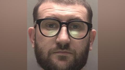 A police mugshot of a man in his early 30s, who has short brown hair and a brown beard. He is wearing black framed glasses and is looking into the camera with a bemused expression. 