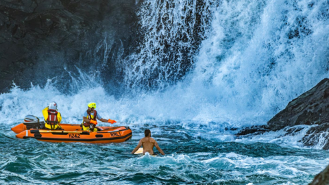The picture shows a rescue scene with an orange inflatable RNLI rescue boat marked with "Y242" and "RNLI", two rescue personnel aboard the boat, wearing helmets and life jackets and a person in the water, on a surfboard.
Water cascades down rocky cliffs into rough, churning water.
