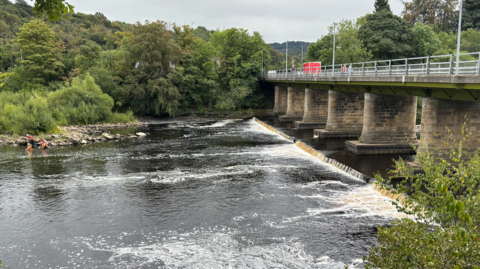 A view of Wylam Bridge, a bridge that crosses the River Tyne. Police and Mountain Rescue teams in hard hats and dry suits are searching the water below the bridge.