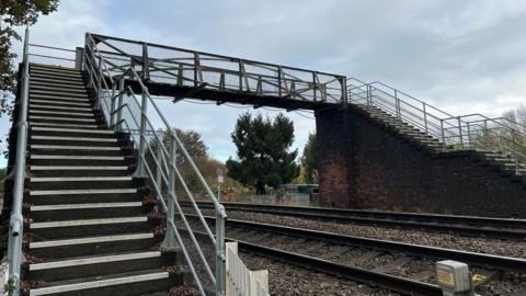 A pedestrian footbridge crosses over a set of railway tracks. The bridge has metal railings and mesh sides, with staircases on both ends one made of concrete steps with metal handrails and the other supported by a brick structure. The railway tracks run horizontally beneath the bridge.
