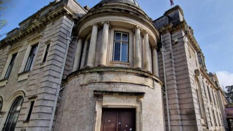 The image is of the rotunda on the white stone Torbay council building. I has classical columns around a first floor window and brown double doors on the ground floor.