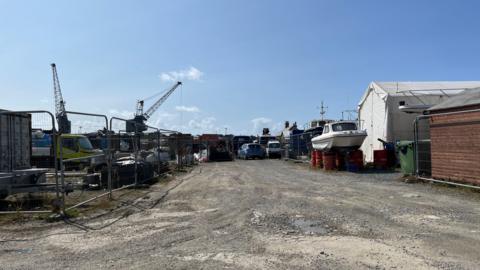 Griffiths Yard - part of it is behind barriers, there is a gravel roadway in the middle and buildings on the right with a boat and some cars