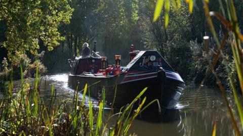The Coventry canal in Fradley, Lichfield. A man steers a barge through the water on a sunny day. The embankment is lined with trees and greenery and the sun bounces off the water.