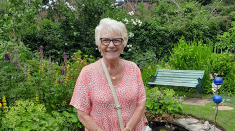 A smiling woman with silver hair and black glasses, wearing a pink spotted dress, stands in a garden