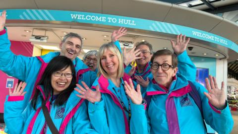 Three women and three men smile and hold up their arms in celebratory fashion in front of "welcome to Hull" signs at a railway station. They are wearing bright blue and pink jackets. 