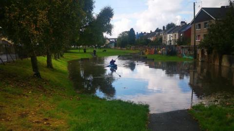 A kayaker paddles through floodwater at a park in Plymouth. The large volume of water is covering a chunk of the park. Green trees line the side of the green grass. Several homes are overlooking the park.