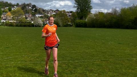 A woman wearing an orange t-shirt and running shorts is running across a grass recreational ground. 