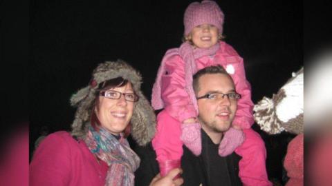 A woman wearing a winter hat with rectangular glasses, a scarf and pink jumper smiling into the camera next to a man with short brown hair also wearing glasses. A young girl wearing all pink including a pink hat is sat on his shoulders smiling.