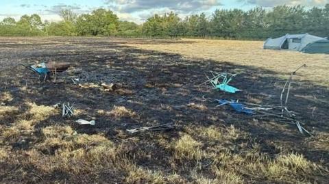Scorched grassland with burnt remnants of camping chairs and a portable barbecue. A wheelbarrow stands next to the barbecue. A green tent is nearby in the field.tent