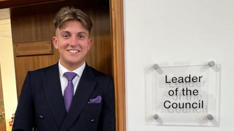 George Finch leans against the door to his office with a sign reading "leader of the council". He is wearing a dark suit with white shirt, purple tie and a purple pocket square in his jacket breast pocket. 