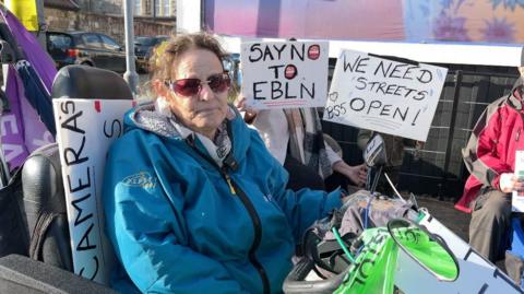 Melissa Topping is sat in a wheelchair and is surrounded by people holding placards, which say "say no to EBLN" and "we need streets open".