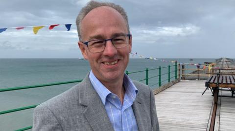 Tim Baker wearing glasses, a blue shirt and grey suit jacket, smiling while standing on the Queen's Pier in Ramsey on a cloudy day.