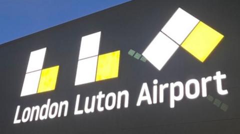 The front of London Luton Airport. A large black building with a large illuminated LLA and "London Luton Airport" written on it in white font.