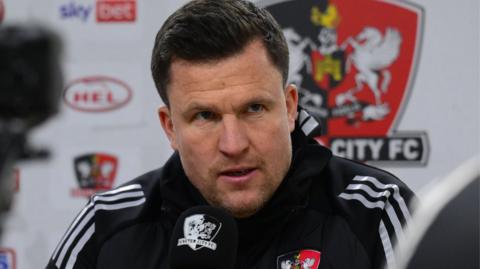 Exeter manager Gary Caldwell conducting a press conference. He has dark hair and is wearing a dark training jacket with the club's badge on.