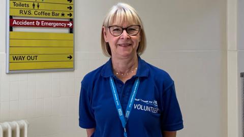 Head and shoulder shot of a middle aged woman, Anne Render. She has blonde bobbed hair with a fringe, is wearing glasses and is smiling. She is wearing a dark blue polo shirt emblazoned with "volunteer" and a blue NHS lanyard. Behind her is a white tiled wall, with a yellow directions signboard to her right.