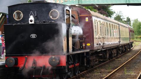 A black steam engine at a station connecting up to three burgundy-coloured carriages. 