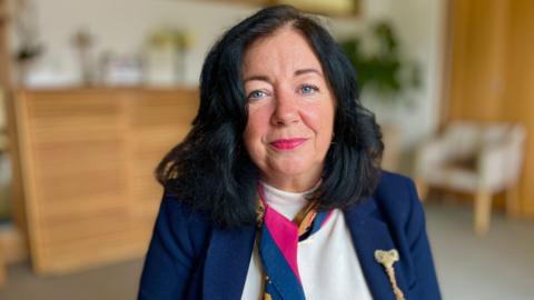 Anne-Marie Hoare looking into camera in a room at the Arthur Rank Hospice. She has long, dark hair and is wearing a navy suit, white jumper and pink and navy scarf.