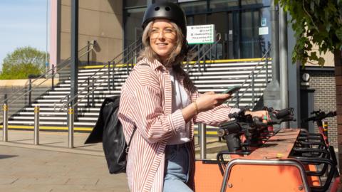A woman, holding a phone, wearing a helmet and standing by a group of e-scooters in a bike rack. She is wearing a red and white striped top, blue jeans and white top. She is looking towards the left and is wearing glasses. Steps to a railway station are behind her. 