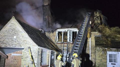 Fire crews tackling a roof fire. Roof is gutted. two staff members in protective clothign hold a ladder. 