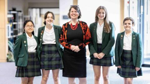 Principal Daniele Harford-Fox wearing a black dress and floral cardigan. She has short dark hair. Two female students are standing either side of her. 
