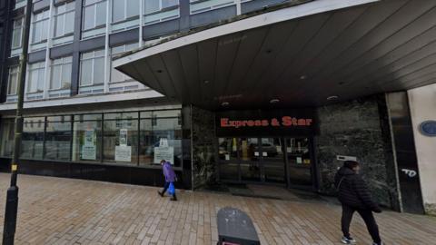 A google streetview picture showing the outside of the former Express and Star newspaper office. There is a person wearing a purple coat and carrying a blue plastic bag walking past the window and a person in a black padded coat standing on the opposite side of the pavement 