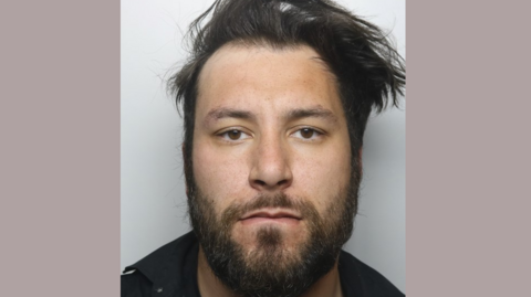 A police custody image of a man with longish black hair, a black beard and brown eyes. He is wearing a dark top. 
