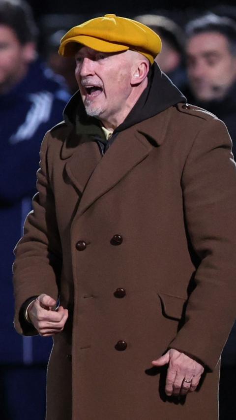 Ian Holloway in a brown coat and yellow hat with his mouth open.