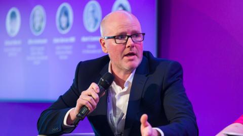 Mark Cramman has glasses, a white shirt and dark suit and is holding a microphone. There is a purple background.