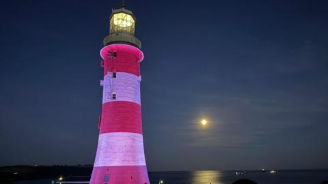 A picture of the lighthouse on Plymouth Hoe. It is red and white in colour, and there is the moon in the distance.