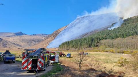 Fire engines parked in front of hills that are covered in smoke