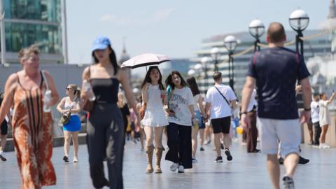 People walk through London in a heatwave, one carrying a bottle of water and another using an umbrella as a sunshade.