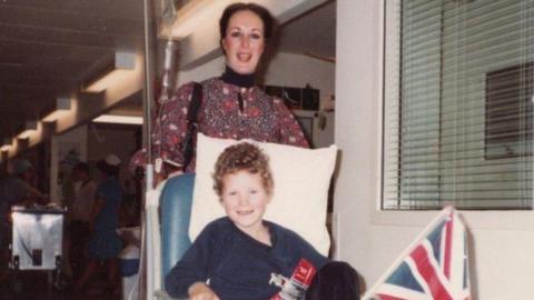 Antonya Cooper stands behind her young son Hamish who sits in a hospital chair.