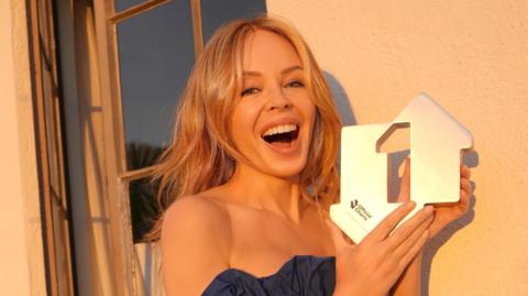Kylie Minogue smiling and holding her Official Charts number one trophy