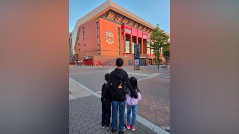 Three young children have their backs turned to the camera as they stand arm in arm looking on at the Anfield stadium.