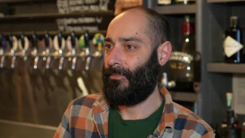 A man with a beard and short hair is inside a bar, with rows of beer taps and chalkboard menus visible in the background.