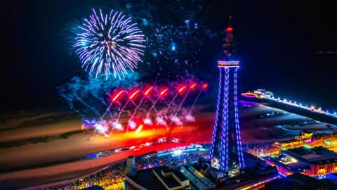A wide view of Blackpool's promenade at night time. There are crowds gathered in front Blackpool Tower to watch the fireworks.