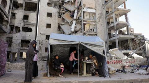 Two women walk past a makeshift tent with three people inside, with destroyed buildings stand in the background