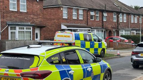 Two police vehicles sit on a narrow residential street. Police tape can be seen ahead, indicating a cordon. Other parked cars are also visible.