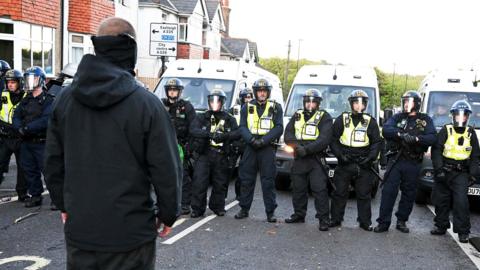 Man wearing a mask and wearing all black and stands in the road with his back to camera. In front of him is a long line of police officers wearing riot gear
